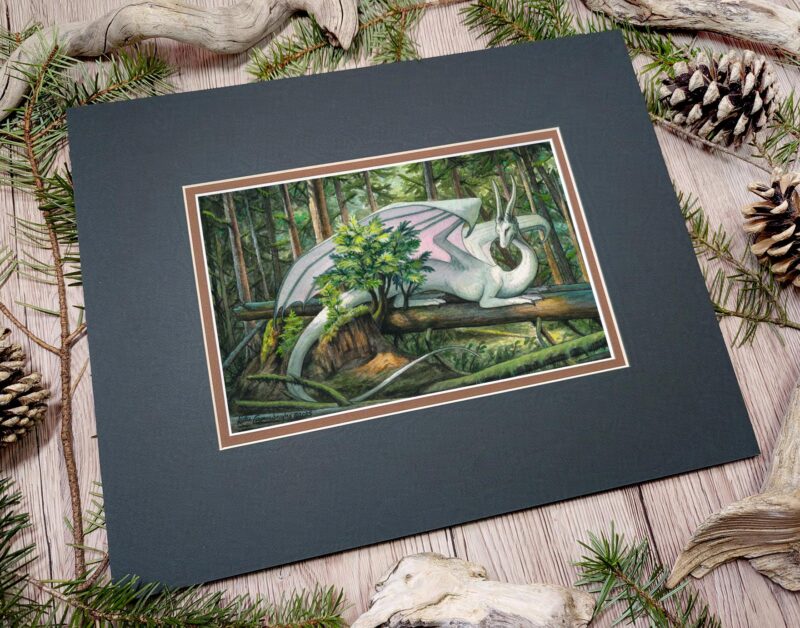 The matted painting sitting on a table with pine cones and wood sticks. A white dragon laying on fallen trees in the forest.