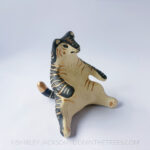 A ceramic tiger sculpture done with a cream white clay and black underglaze with sgraffito markings all over it's body. This shows the right side of the piece. The tiger is in a seated-up position almost similar to a human sitting down with their legs splayed out in front of them.