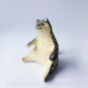 A ceramic tiger sculpture done with a cream white clay and black underglaze with sgraffito markings all over it's body. This shows the left side of the piece. The tiger is in a seated-up position almost similar to a human sitting down with their legs splayed out in front of them.