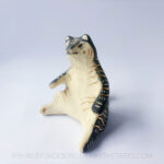 A ceramic tiger sculpture done with a cream white clay and black underglaze with sgraffito markings all over it's body. This shows the left side of the piece. The tiger is in a seated-up position almost similar to a human sitting down with their legs splayed out in front of them.