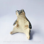 A ceramic tiger sculpture done with a cream white clay and black underglaze with sgraffito markings all over it's body. This shows the front of the piece. The tiger is in a seated-up position almost similar to a human sitting down with their legs splayed out in front of them.