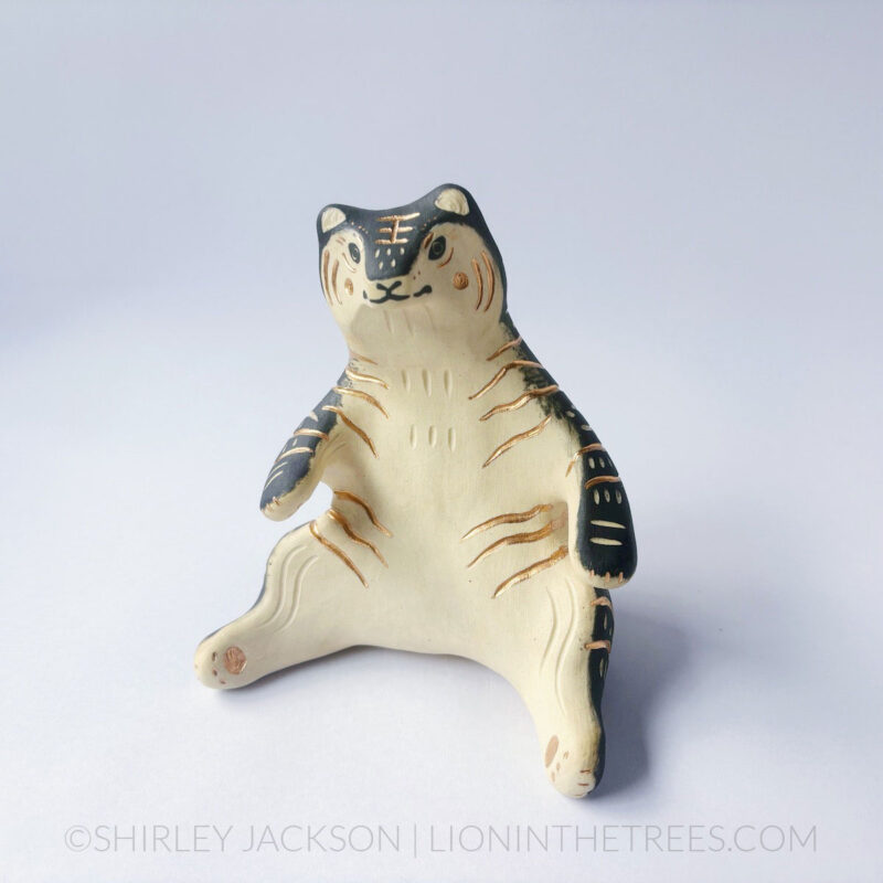 A ceramic tiger sculpture done with a cream white clay and black underglaze with sgraffito markings all over it's body. This shows the front of the piece. The tiger is in a seated-up position almost similar to a human sitting down with their legs splayed out in front of them.