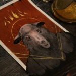 A painting of a sheep with red eyes. The background is solid red with black drips at the top. Above the sheep's head is a broken gold crown. A small gold star and four gold lines burst out from the sheep's chest.
