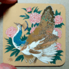 Painting on a square coaster of a pied peahen surrounded by pink peonies, photographed with the artist's thumb and index finger for scale.