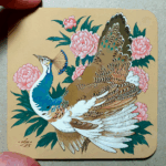 Painting on a square coaster of a pied peahen surrounded by pink peonies, photographed with the artist's thumb and index finger for scale.