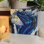 A painting of a blue dragon on a canvas sitting in a table surrounded by candles and crystals with plants in the background