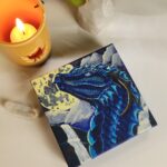 A painting of a blue dragon on a canvas sitting in a table surrounded by candles and crystals with plants in the background