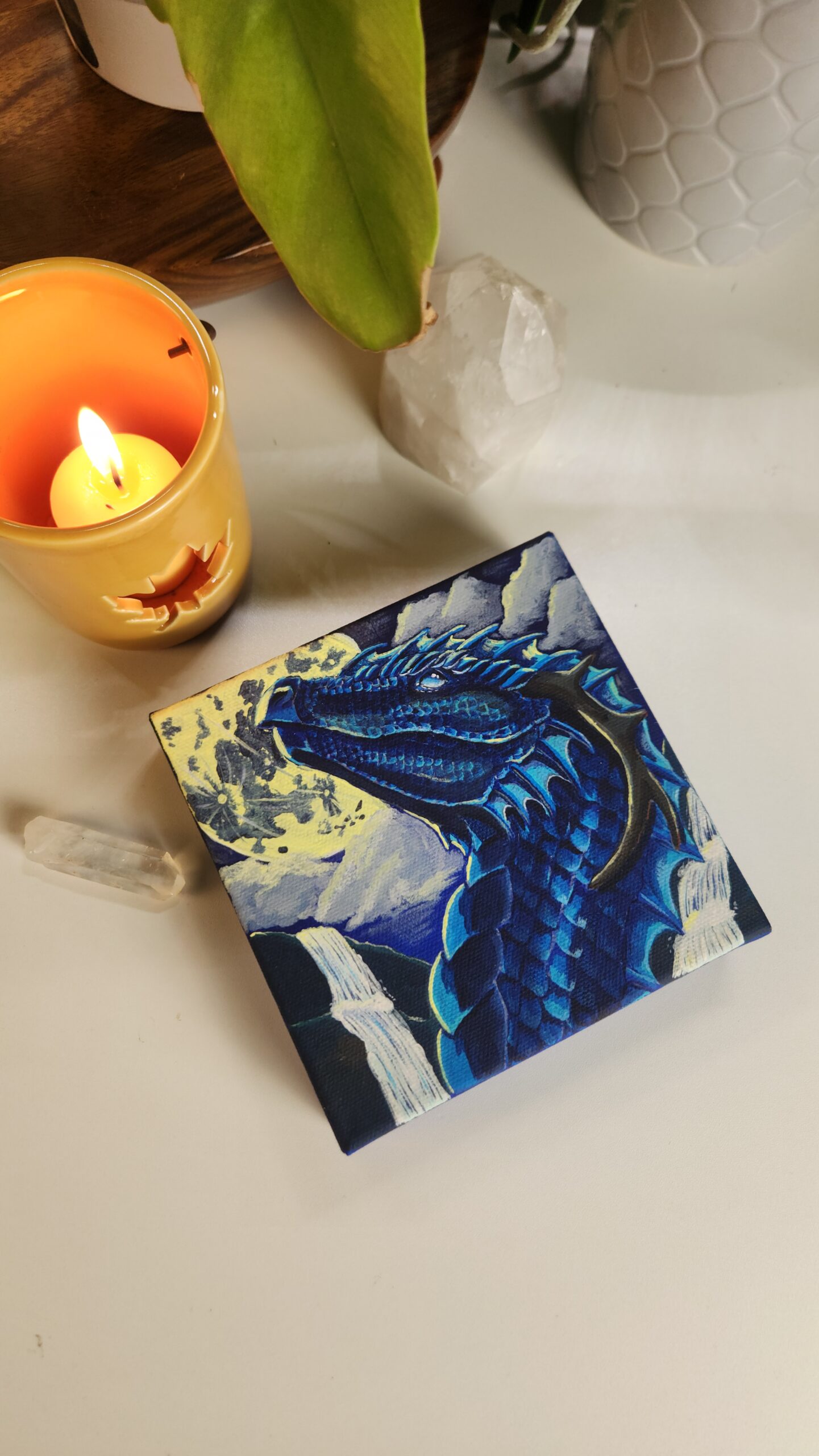 A painting of a blue dragon on a canvas sitting in a table surrounded by candles and crystals with plants in the background