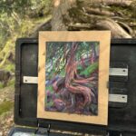 A plein-air painting of a twisted cedar tree with colorful roots and dappled forest light, displayed on an easel in a wooded lakeshore setting.