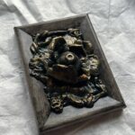 A photo of a sculpture of an eyeball flower surrounded by branches and flora. The sculpture is dark in color, with green patina effect in some crevices and gold highlighting the textures.