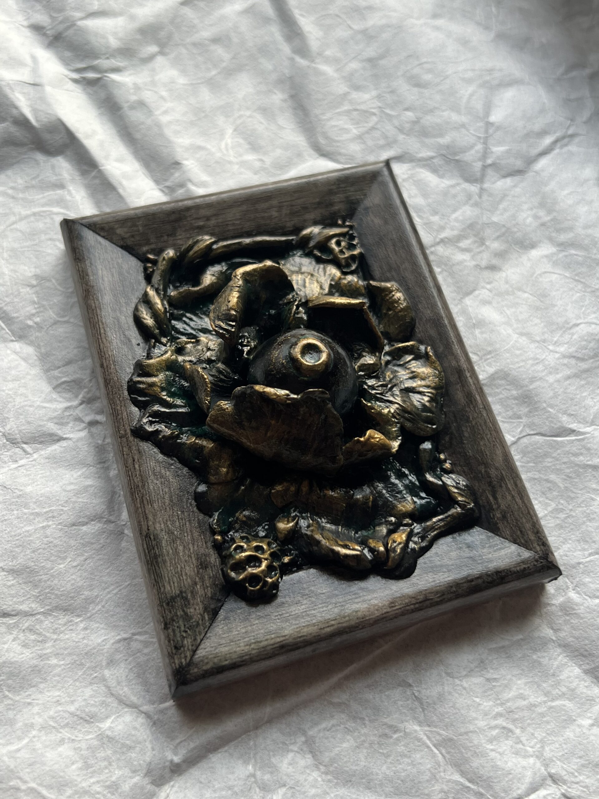 A photo of a sculpture of an eyeball flower surrounded by branches and flora. The sculpture is dark in color, with green patina effect in some crevices and gold highlighting the textures.