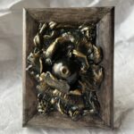 A photo of a sculpture of an eyeball flower surrounded by branches and flora. The sculpture is dark in color, with green patina effect in some crevices and gold highlighting the textures.