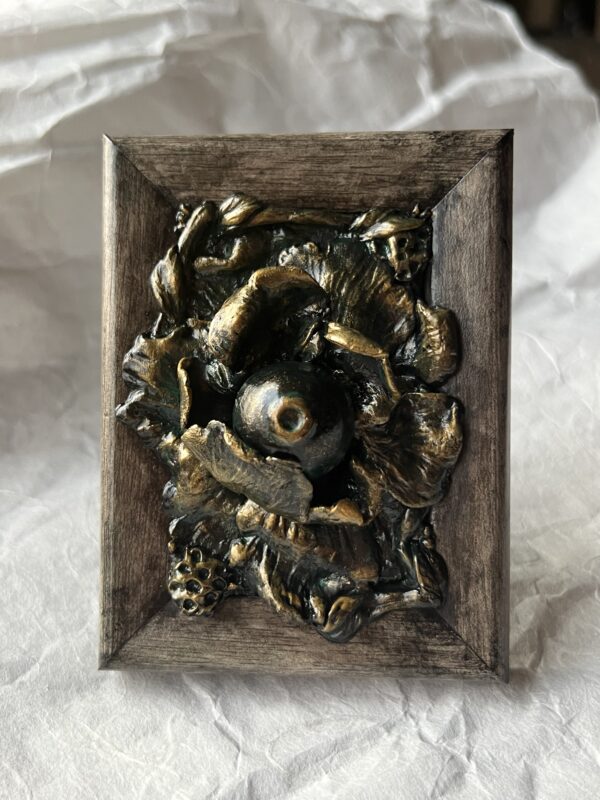 A photo of a sculpture of an eyeball flower surrounded by branches and flora. The sculpture is dark in color, with green patina effect in some crevices and gold highlighting the textures.