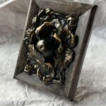 A photo of a sculpture of an eyeball flower surrounded by branches and flora. The sculpture is dark in color, with green patina effect in some crevices and gold highlighting the textures.