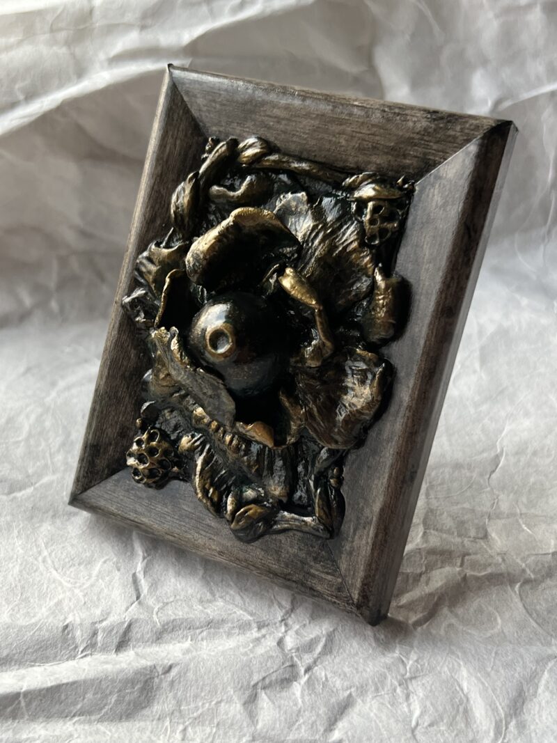 A photo of a sculpture of an eyeball flower surrounded by branches and flora. The sculpture is dark in color, with green patina effect in some crevices and gold highlighting the textures.