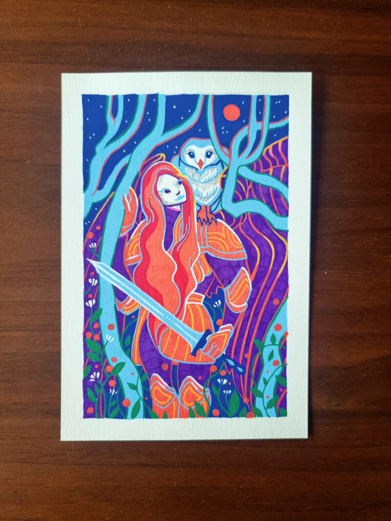 An artwork named "Interlude" lies on top of a wooden table. It depicts a woman in armor, holding a sword looking up to the moon with an owl perched on her left shoulder. They are framed by trees and flowers with a mountain & sky behind them. The artwork is colored predominantly in blue, orange and violet posca pen with a white border.