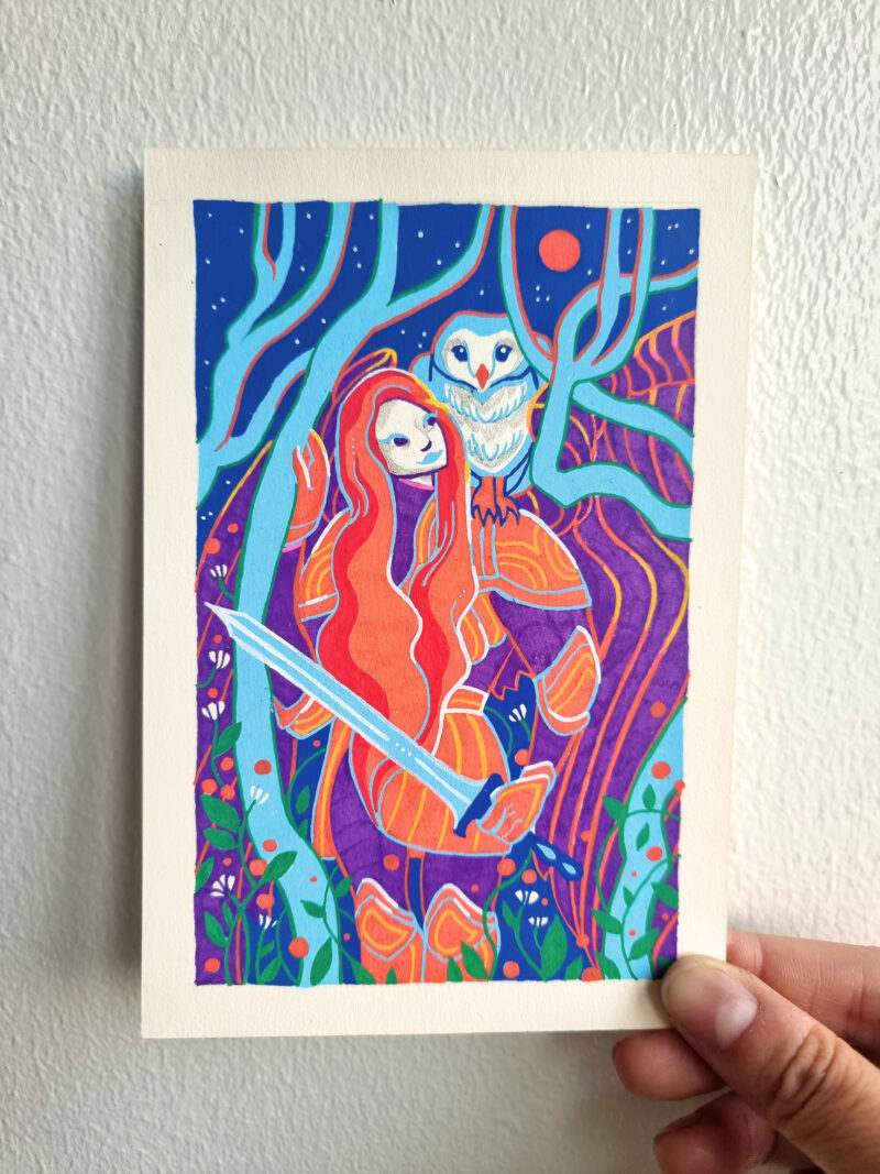 An artwork named "Interlude" is held by the artist's right hand in front of a white wall. It depicts a woman in armor, holding a sword looking up to the moon with an owl perched on her left shoulder. They are framed by trees and flowers with a mountain & sky behind them. The artwork is colored predominantly in blue, orange and violet posca pen with a white border.
