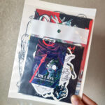 A pack of postcards & stickers of the artist's merch that will accompany the purchase of "Interlude" artwork. It is held up by the artist's hand on grey wall background.