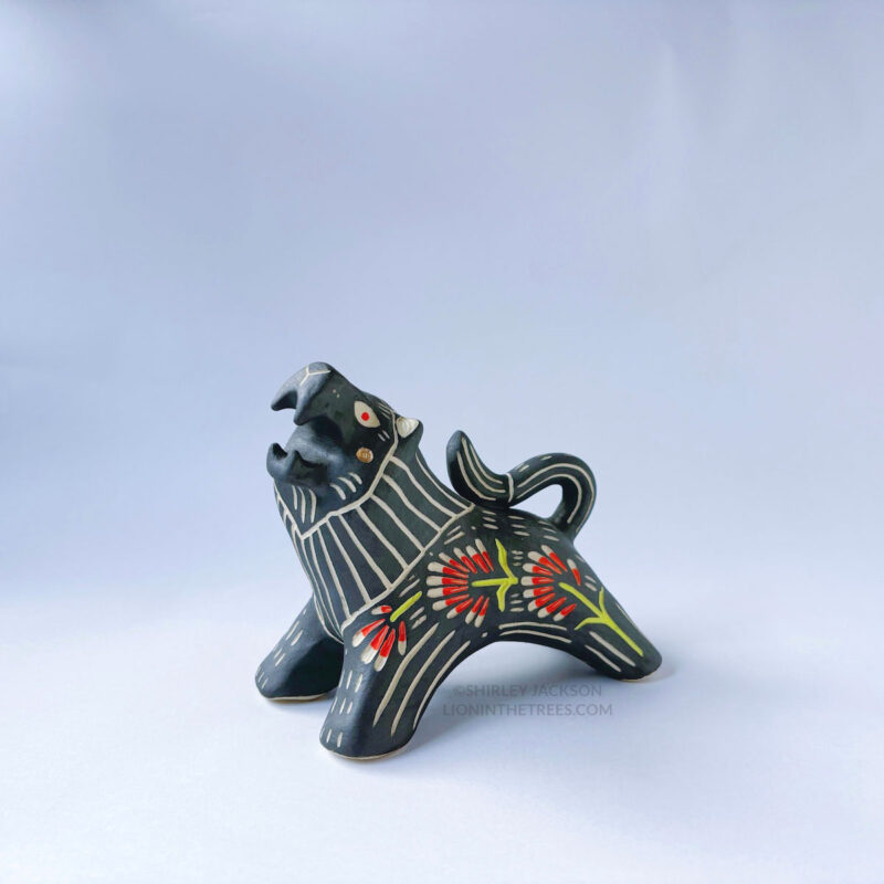 A ceramic lion sculpture with it's mouth open done with black underglaze. The piece features sgraffito Prairie Fire, a plant that looks like a fire-y paintbrush, carved onto the lion's side done with red and green underglazes.