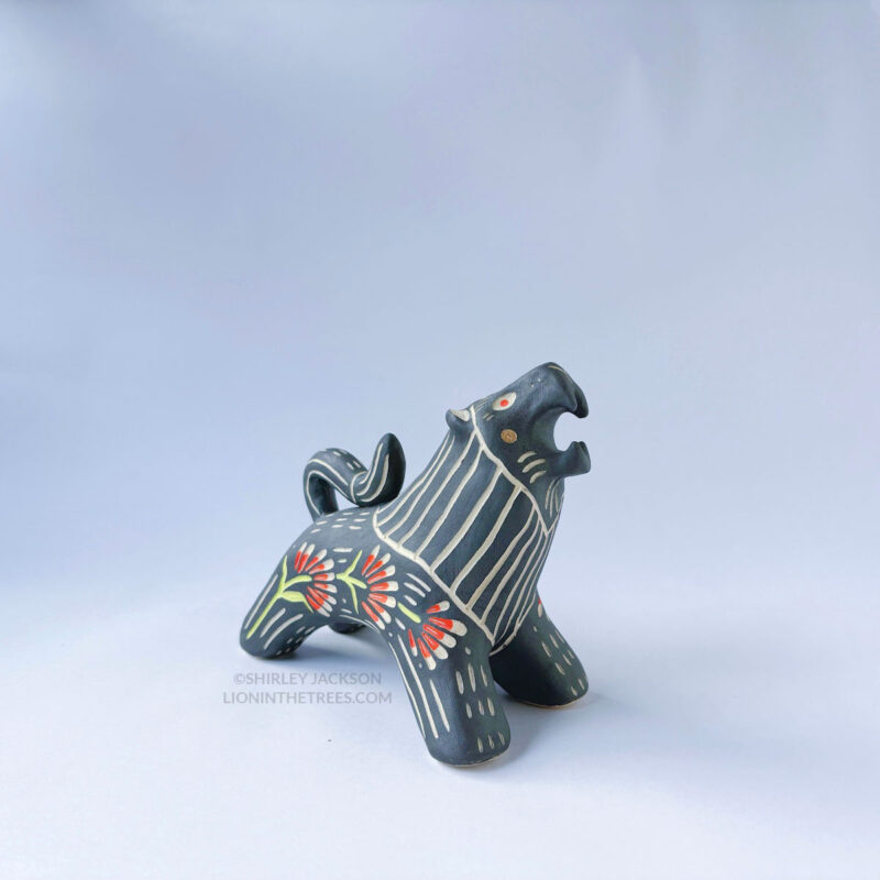 A ceramic lion sculpture with it's mouth open done with black underglaze. The piece features sgraffito Prairie Fire, a plant that looks like a fire-y paintbrush, carved onto the lion's side done with red and green underglazes.