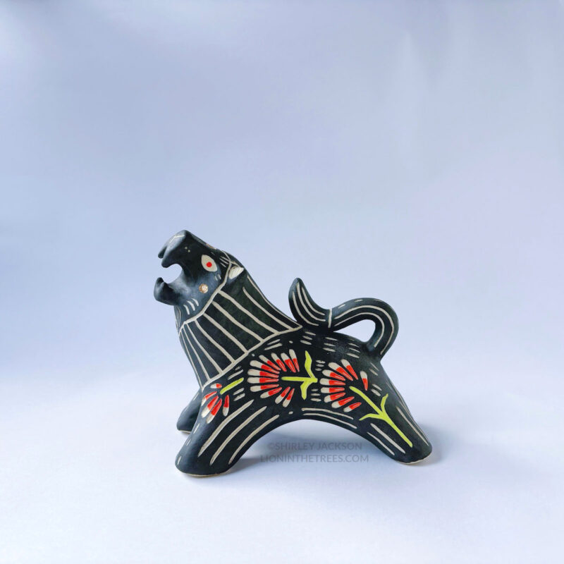 A ceramic lion sculpture with it's mouth open done with black underglaze. The piece features sgraffito Prairie Fire, a plant that looks like a fire-y paintbrush, carved onto the lion's side done with red and green underglazes.