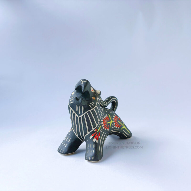 A ceramic lion sculpture with it's mouth open done with black underglaze. The piece features sgraffito Prairie Fire, a plant that looks like a fire-y paintbrush, carved onto the lion's side done with red and green underglazes.