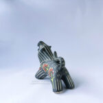 A ceramic lion sculpture with it's mouth open done with black underglaze. The piece features sgraffito Prairie Fire, a plant that looks like a fire-y paintbrush, carved onto the lion's side done with red and green underglazes. This image shows the back of the sculpture.