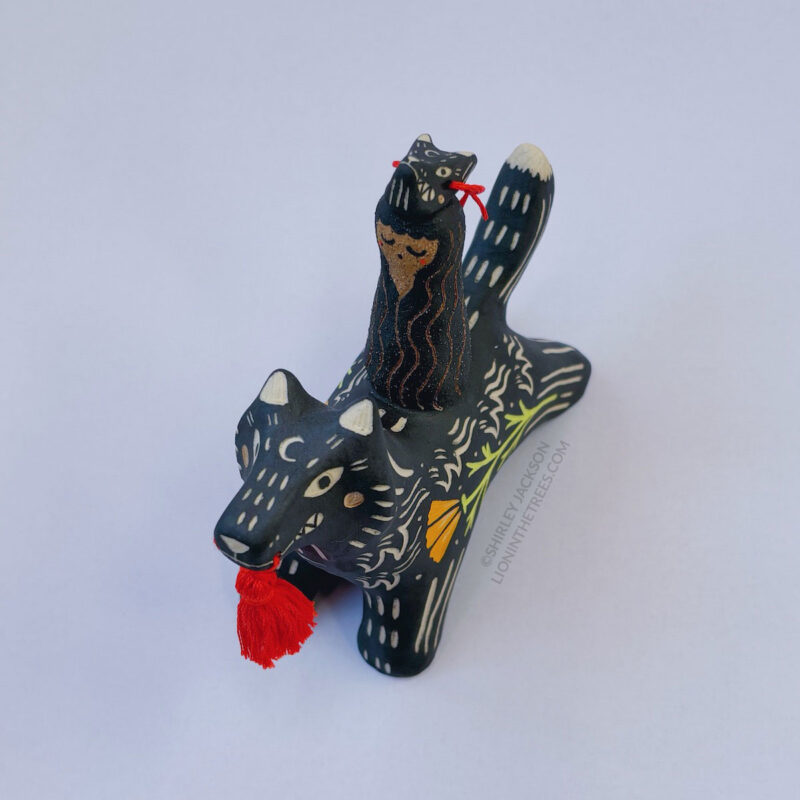 A black ceramic wolf sculpture with a variety of carved, sgraffito marks all along it's body including my signature California Poppy motif on it's side and textures of fur, and a handmade red tassel hanging from it’s mouth. There is a brown coloured woman with long wavy black hair sitting on the back of the wolf with her eyes closed as though she is singing. She has a matching black wolf mask on top of her head. This image is a bird's eye view to show the wolf mask on the head of the woman.