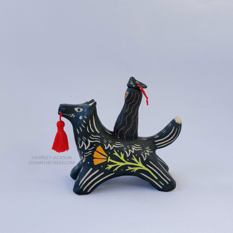 A black ceramic wolf sculpture with a variety of carved, sgraffito marks all along it's body including my signature California Poppy motif on it's side and textures of fur, and a handmade red tassel hanging from it’s mouth. There is a brown coloured woman with long wavy black hair sitting on the back of the wolf with her eyes closed as though she is singing. She has a matching black wolf mask on top of her head.