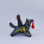 A black ceramic wolf sculpture with a variety of carved, sgraffito marks all along it's body including my signature California Poppy motif on it's side and textures of fur, and a handmade red tassel hanging from it’s mouth. There is a brown coloured woman with long wavy black hair sitting on the back of the wolf with her eyes closed as though she is singing. She has a matching black wolf mask on top of her head.