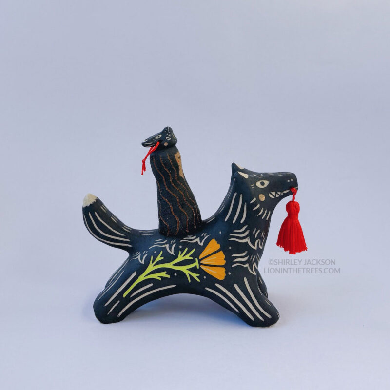 A black ceramic wolf sculpture with a variety of carved, sgraffito marks all along it's body including my signature California Poppy motif on it's side and textures of fur, and a handmade red tassel hanging from it’s mouth. There is a brown coloured woman with long wavy black hair sitting on the back of the wolf with her eyes closed as though she is singing. She has a matching black wolf mask on top of her head.