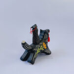 A black ceramic wolf sculpture with a variety of carved, sgraffito marks all along it's body including my signature California Poppy motif on it's side and textures of fur, and a handmade red tassel hanging from it’s mouth. There is a brown coloured woman with long wavy black hair sitting on the back of the wolf with her eyes closed as though she is singing. She has a matching black wolf mask on top of her head.