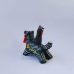 A black ceramic wolf sculpture with a variety of carved, sgraffito marks all along it's body including my signature California Poppy motif on it's side and textures of fur, and a handmade red tassel hanging from it’s mouth. There is a brown coloured woman with long wavy black hair sitting on the back of the wolf with her eyes closed as though she is singing. She has a matching black wolf mask on top of her head.