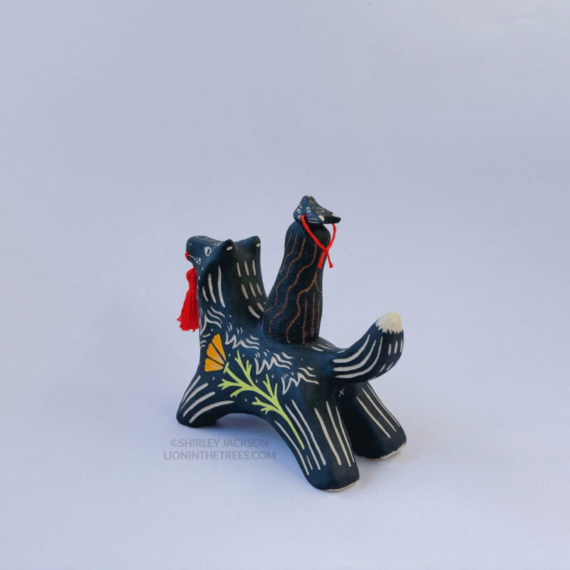 A black ceramic wolf sculpture with a variety of carved, sgraffito marks all along it's body including my signature California Poppy motif on it's side and textures of fur, and a handmade red tassel hanging from it’s mouth. There is a brown coloured woman with long wavy black hair sitting on the back of the wolf with her eyes closed as though she is singing. She has a matching black wolf mask on top of her head.
