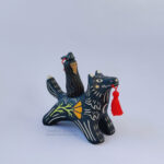 A black ceramic wolf sculpture with a variety of carved, sgraffito marks all along it's body including my signature California Poppy motif on it's side and textures of fur, and a handmade red tassel hanging from it’s mouth. There is a brown coloured woman with long wavy black hair sitting on the back of the wolf with her eyes closed as though she is singing. She has a matching black wolf mask on top of her head.