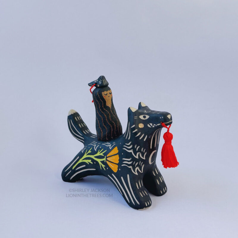 A black ceramic wolf sculpture with a variety of carved, sgraffito marks all along it's body including my signature California Poppy motif on it's side and textures of fur, and a handmade red tassel hanging from it’s mouth. There is a brown coloured woman with long wavy black hair sitting on the back of the wolf with her eyes closed as though she is singing. She has a matching black wolf mask on top of her head.