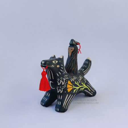 A black ceramic wolf sculpture with a variety of carved, sgraffito marks all along it's body including my signature California Poppy motif on it's side and textures of fur, and a handmade red tassel hanging from it’s mouth. There is a brown coloured woman with long wavy black hair sitting on the back of the wolf with her eyes closed as though she is singing. She has a matching black wolf mask on top of her head.