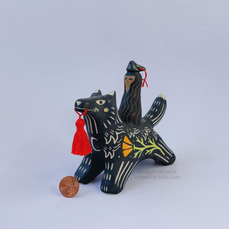 A black ceramic wolf sculpture with a variety of carved, sgraffito marks all along it's body including my signature California Poppy motif on it's side and textures of fur, and a handmade red tassel hanging from it’s mouth. There is a brown coloured woman with long wavy black hair sitting on the back of the wolf with her eyes closed as though she is singing. She has a matching black wolf mask on top of her head. This photo features a penny taken with the sculpture to show scale.