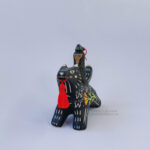 A black ceramic wolf sculpture with a variety of carved, sgraffito marks all along it's body including my signature California Poppy motif on it's side and textures of fur, and a handmade red tassel hanging from it’s mouth. There is a brown coloured woman with long wavy black hair sitting on the back of the wolf with her eyes closed as though she is singing. She has a matching black wolf mask on top of her head.