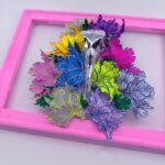 A hand-sculpted silver filigree bird skull nestled in a riot of colourful 3d flowers, hand-stitched to the frame