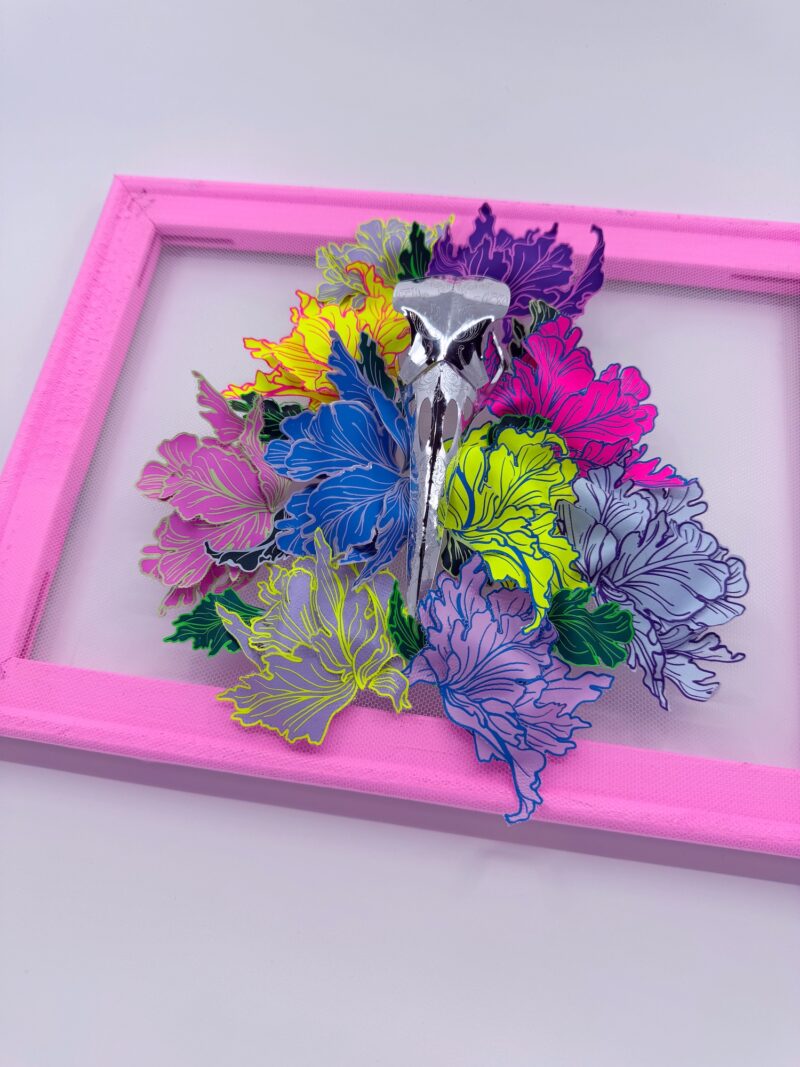 A hand-sculpted silver filigree bird skull nestled in a riot of colourful 3d flowers, hand-stitched to the frame