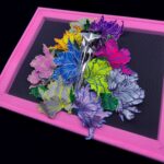 A hand-sculpted silver filigree bird skull nestled in a riot of colourful 3d flowers, hand-stitched to the frame