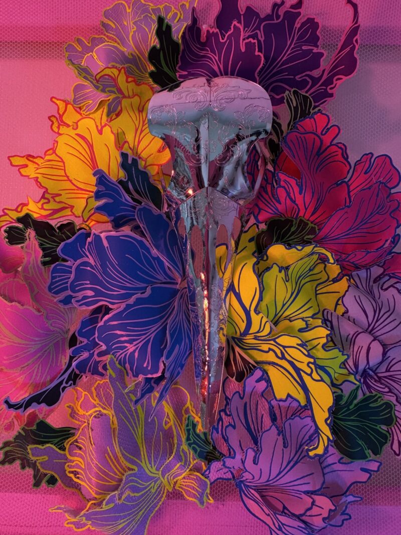 A hand-sculpted silver filigree bird skull nestled in a riot of colourful 3d flowers, hand-stitched to the frame