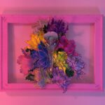 A hand-sculpted silver filigree bird skull nestled in a riot of colourful 3d flowers, hand-stitched to the frame