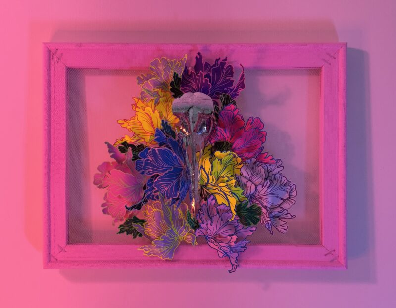 A hand-sculpted silver filigree bird skull nestled in a riot of colourful 3d flowers, hand-stitched to the frame