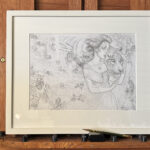 Studio shot of "Strength" drawing framed and matted in white on a mahogany easel. A pencil rests in front of the framed drawing.