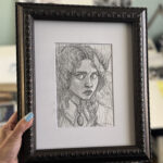 The artist's hand holds framed drawing against studio background for scale.