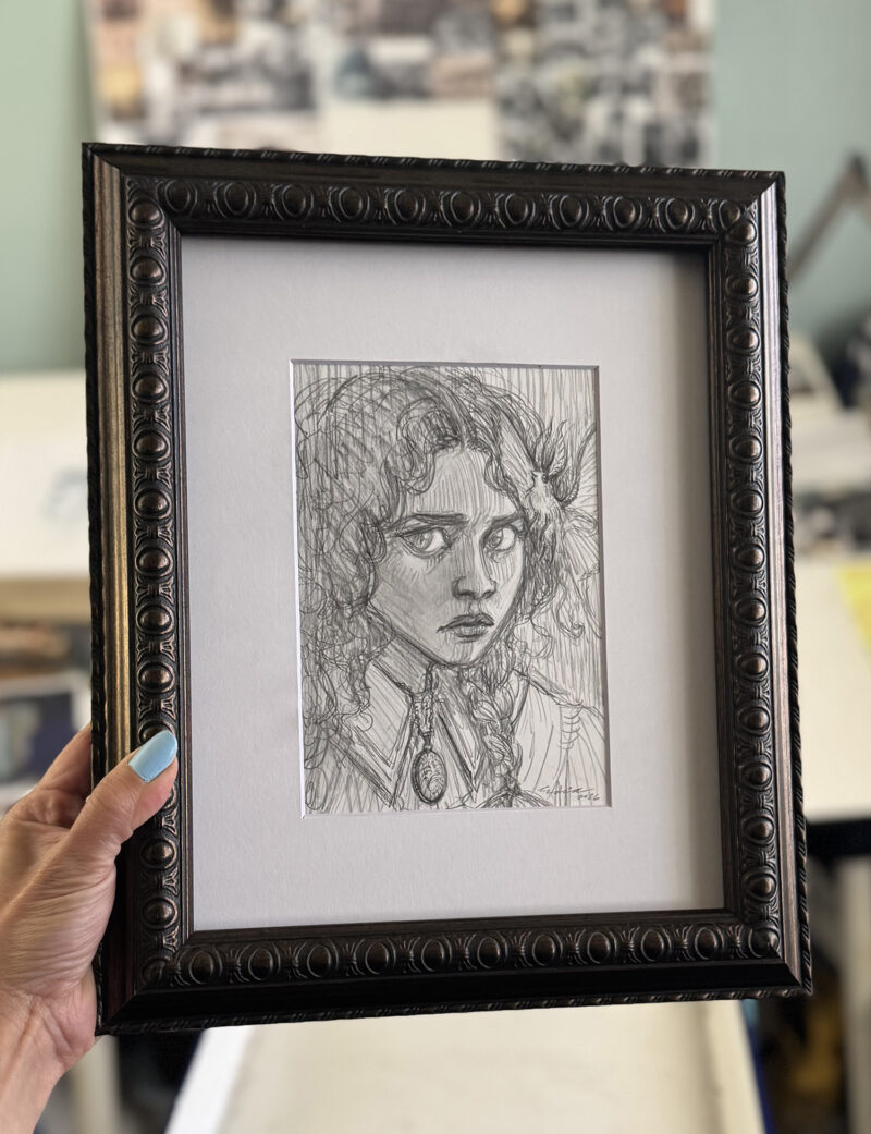 The artist's hand holds framed drawing against studio background for scale.