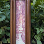 Painting in a narrow frame set on some leaves to display. It is a pink and orange abstract landscape of sunset.