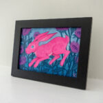 A framed painting of a pink hare surrounded by purple bee balm bergamot flowers.